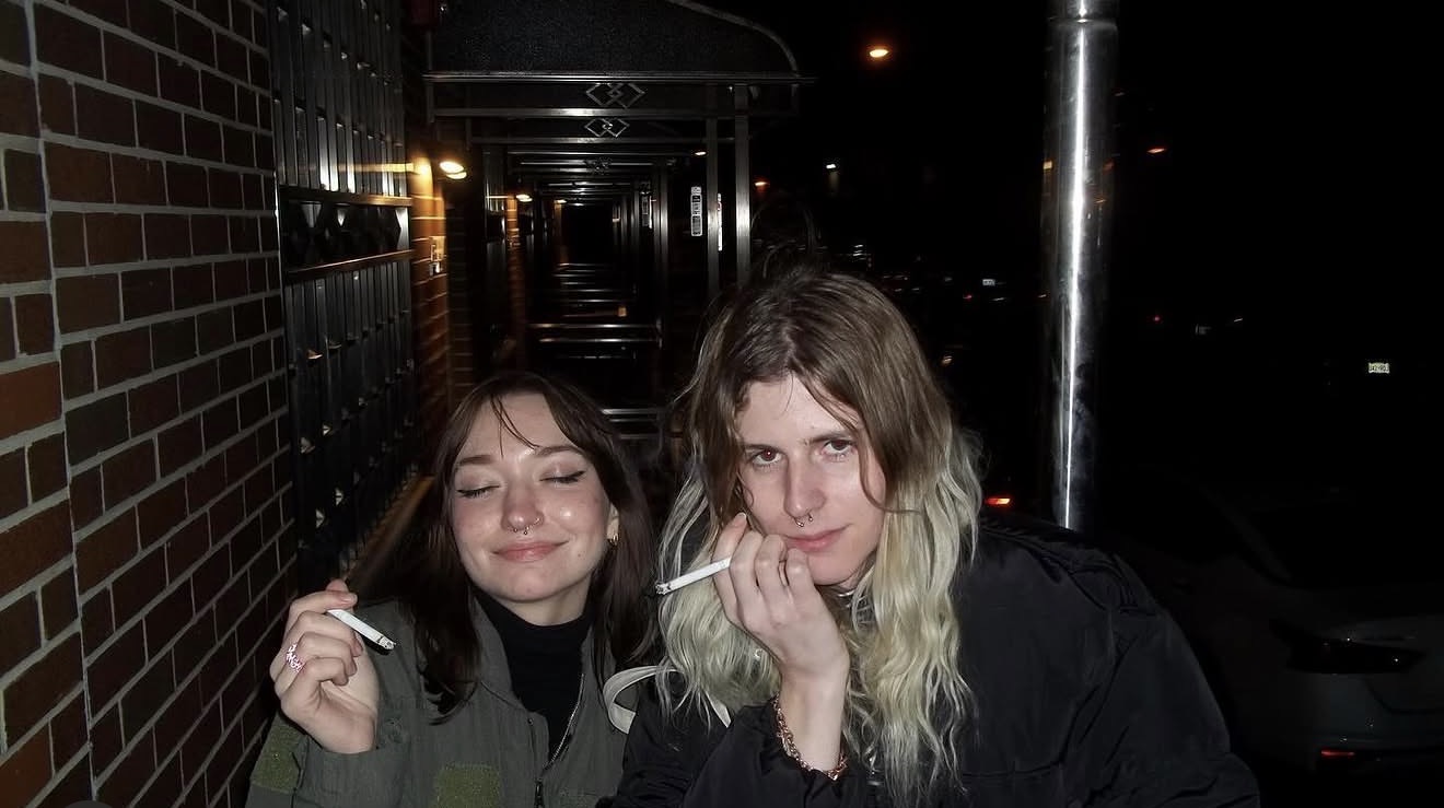 Anna with a friend outside at night, sharing a cigarette on a brick-lined sidewalk